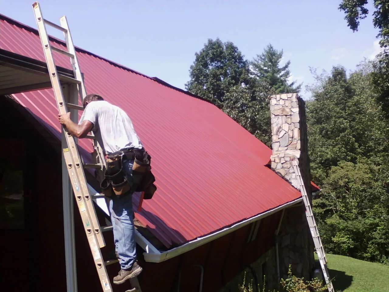 Roofer on a ladder beside a red metal roof and stone chimney — Soffit & Fascia Repair by Roofing Brothers in Kawkawlin