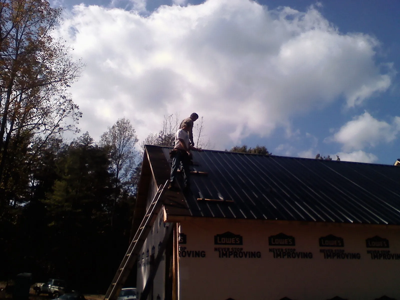 Roofer fastening dark metal panels high against a cloudy sky, from a Roof Inspection job in Kawkawlin