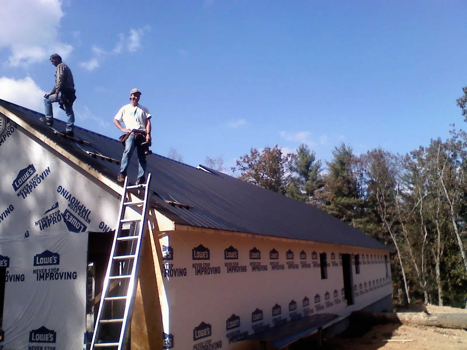 Two roofers standing on a long dark metal roof over a wrapped building in Kawkawlin